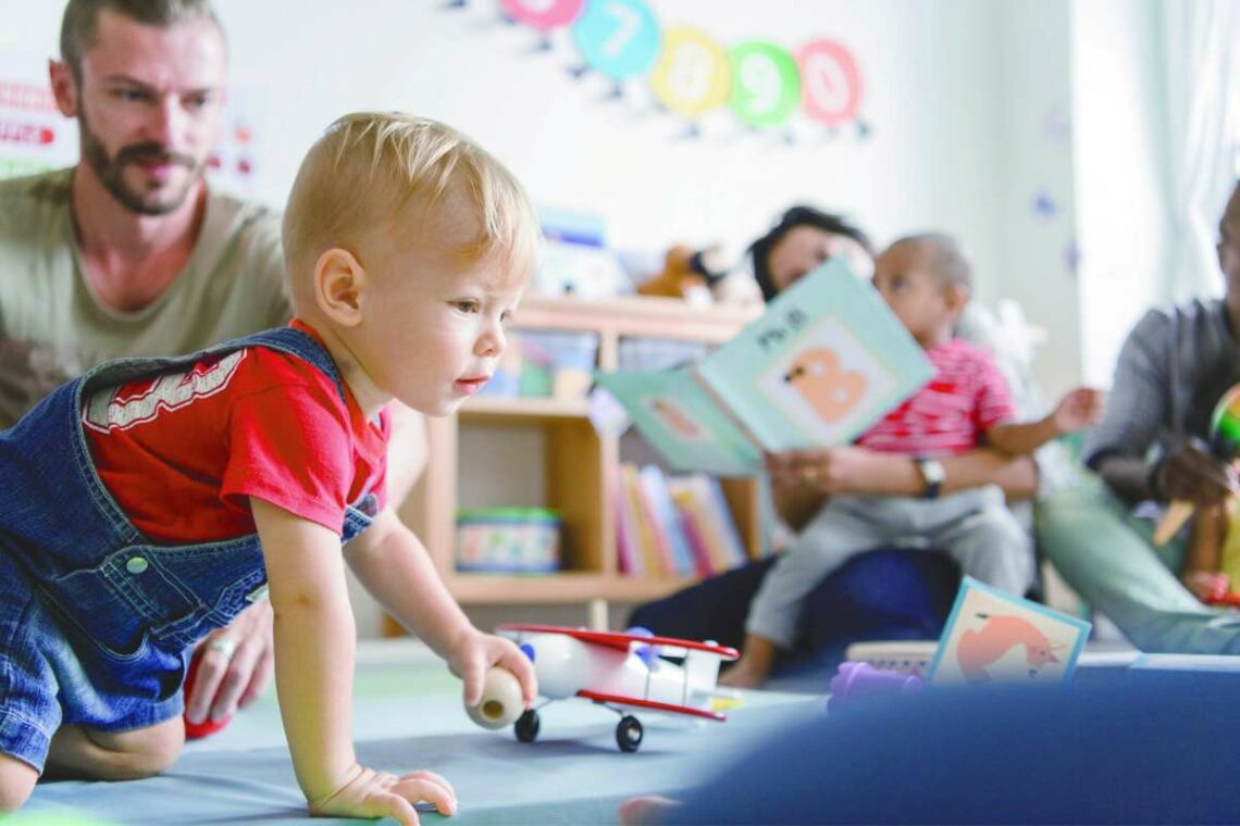 Un papa observe son jeune enfant qui explore à quatre pattes dans un lieu d'accueil enfants-parents, pendant que d'autres parents lisent avec leurs enfants