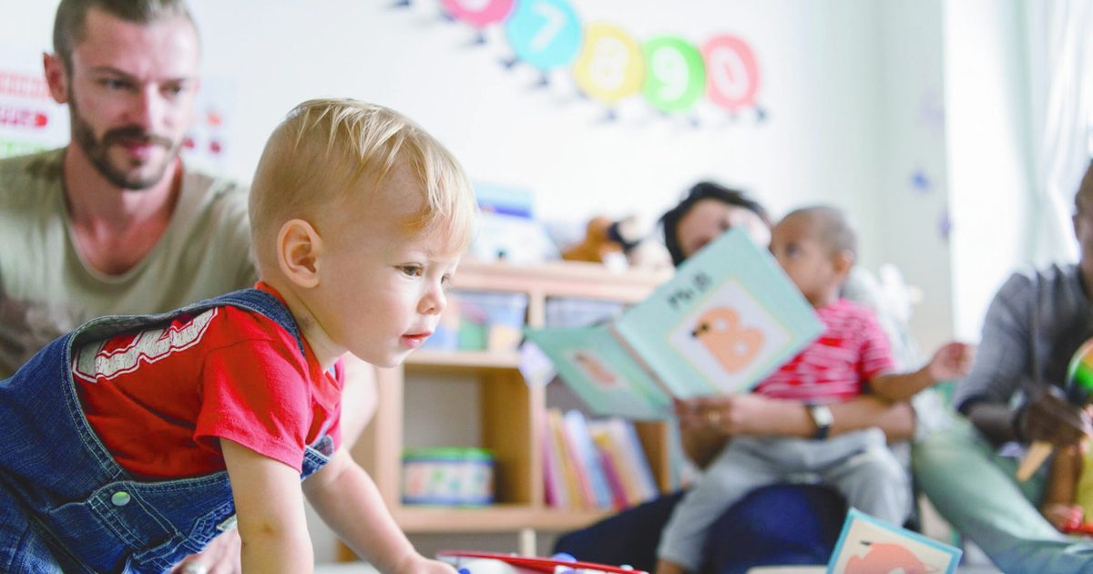 Un papa observe son jeune enfant qui explore à quatre pattes dans un lieu d'accueil enfants-parents, pendant que d'autres parents lisent avec leurs enfants